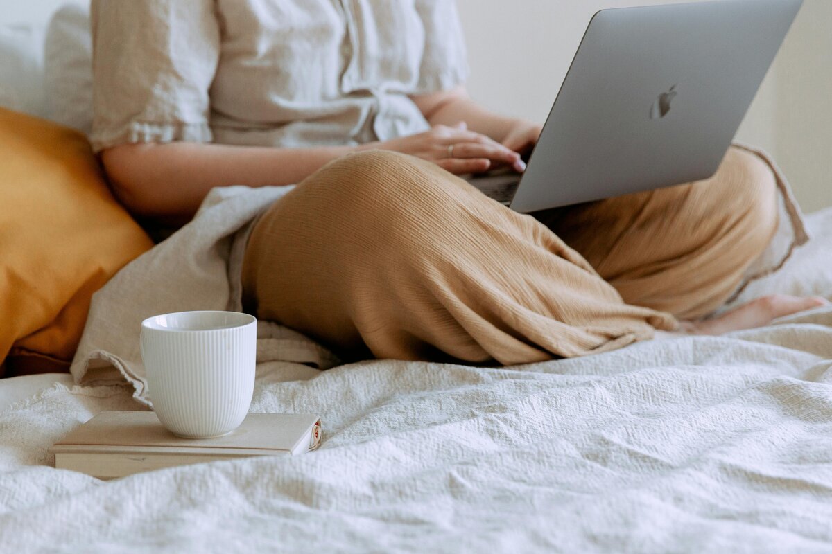 Woman sitting cross legged on a bed with a laptop and coffee, working comfortably from home