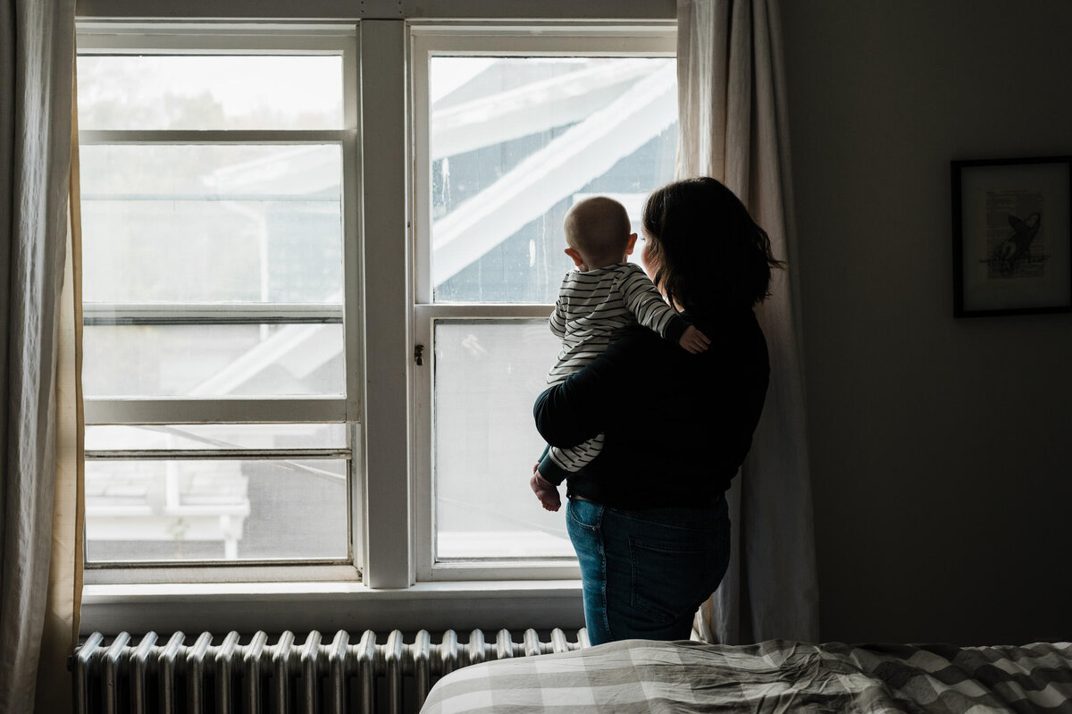 Woman holding baby and looking out bedroom window.