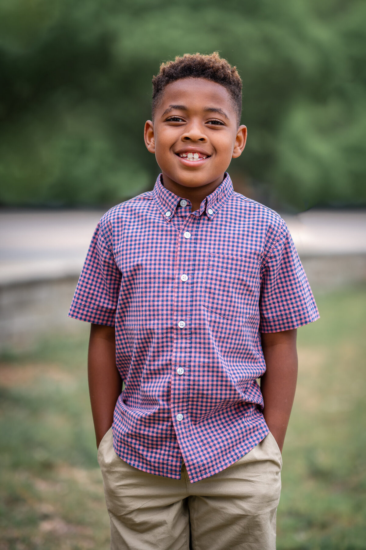 A young boy wearing a plaid button down shirt has his hands in his pocket and is smiling.