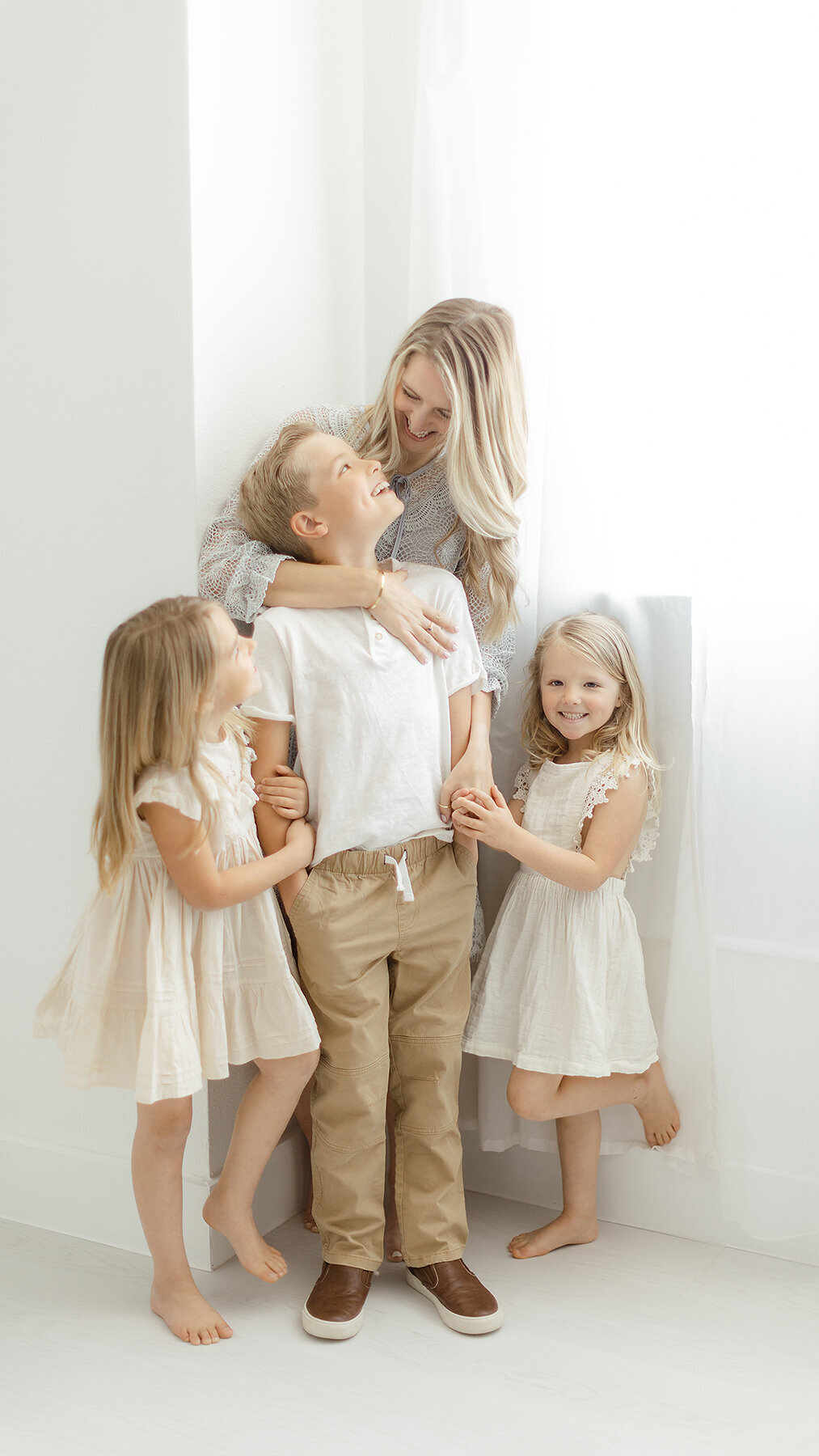A mother and her 3 young children standing by the window of a Dallas photography studio looking an smiling at each other.