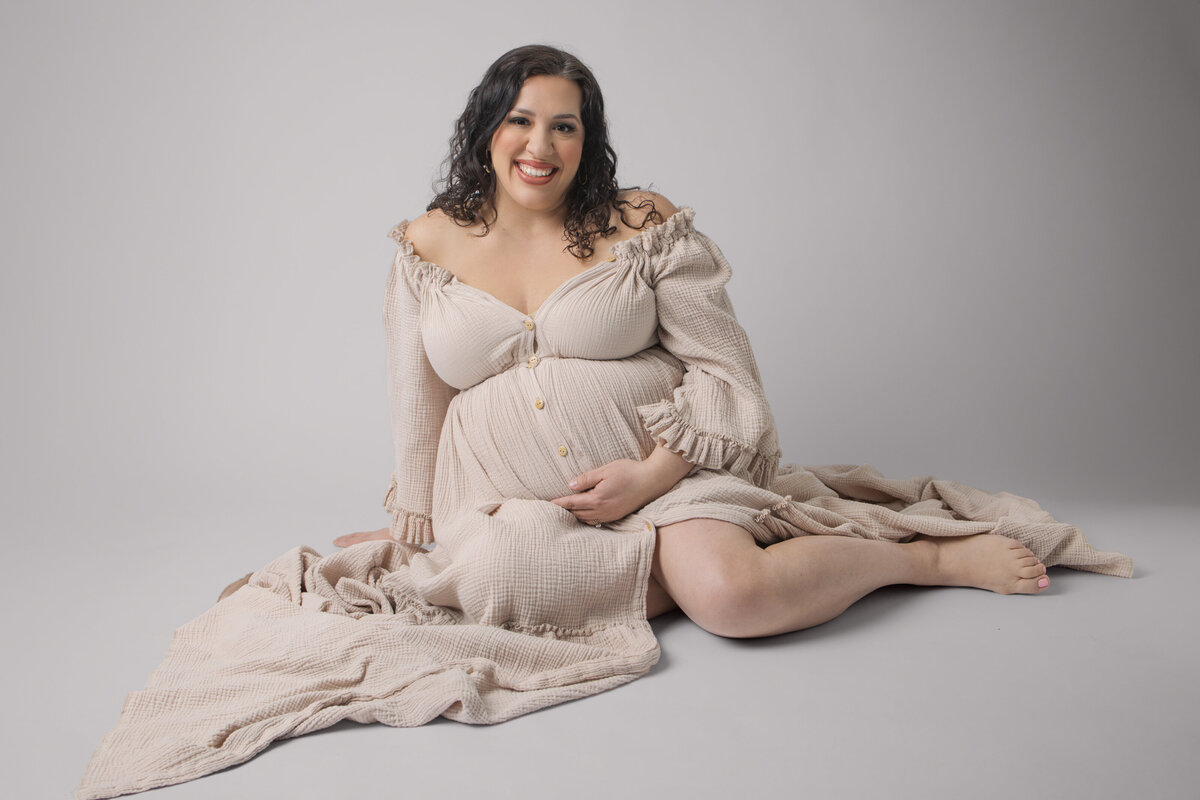 Expectant mother in cream colored dress poses gracefully on floor with flowing fabric during maternity photoshoot near Hackensack, NJ.