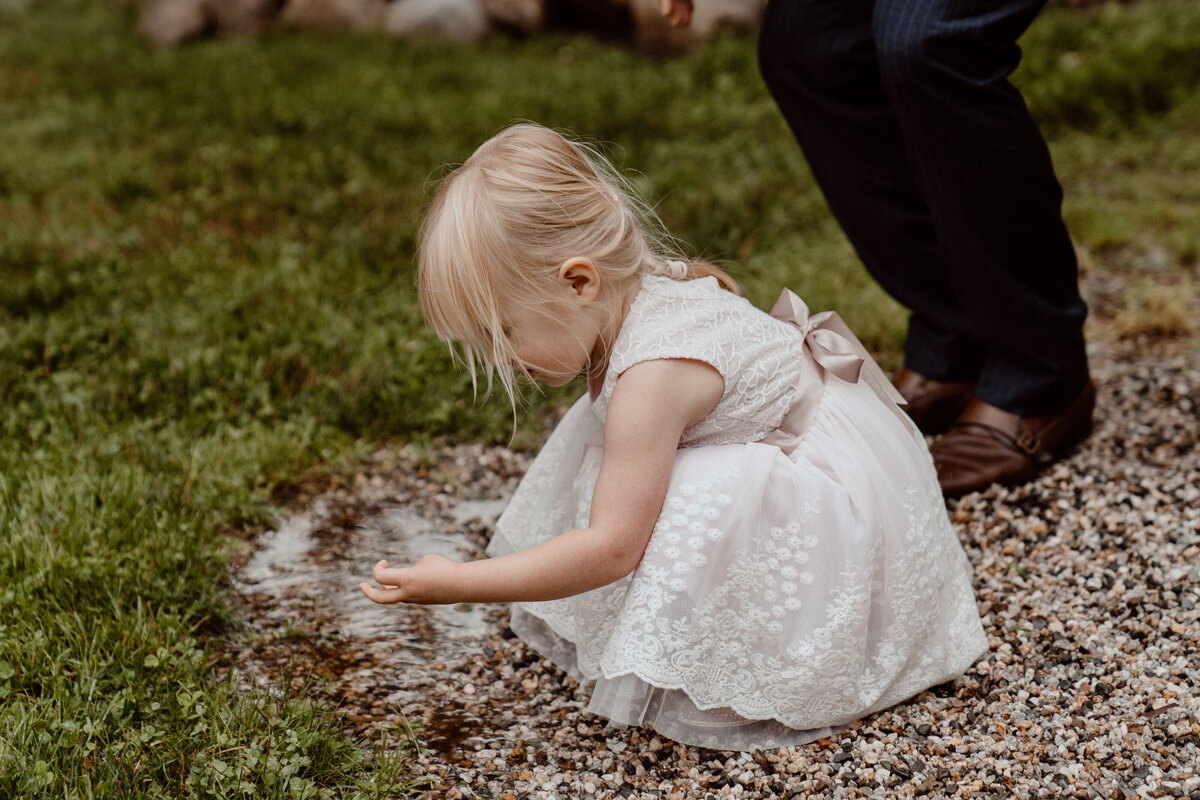 Adorable flower girl in a lace dress plays with water on a gravel path during a wedding ceremony. Captured in a candid, documentary style, the young child, with blonde hair, is engrossed in her exploration, highlighting the innocence and joy of the wedding day.