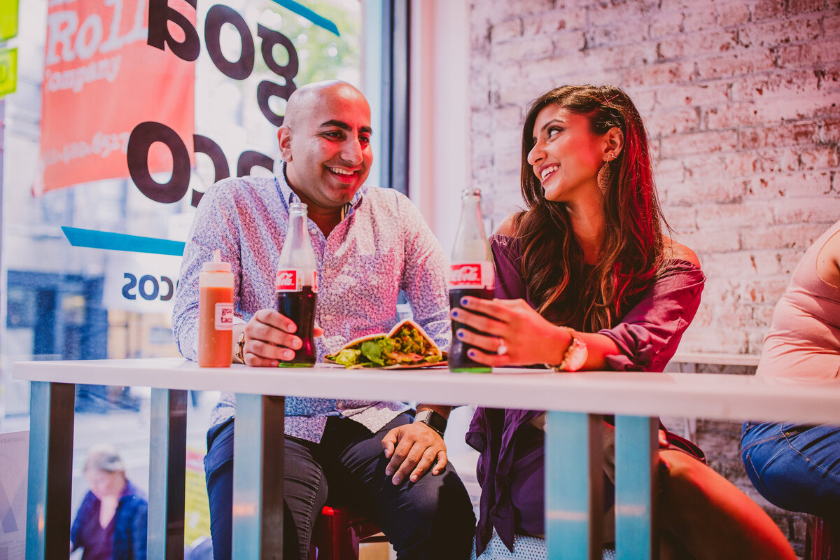 A couple sitting down and talking to each other as they share a taco and coke bottles inside a taqueria on macdougal street in nyc