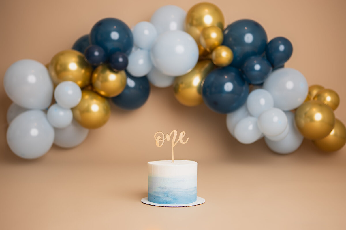 Savage seamless pecan backdrop with light blue navy blue and gold balloons. Cake by Triple L Cakes and cake topper by Etch'd Designs
