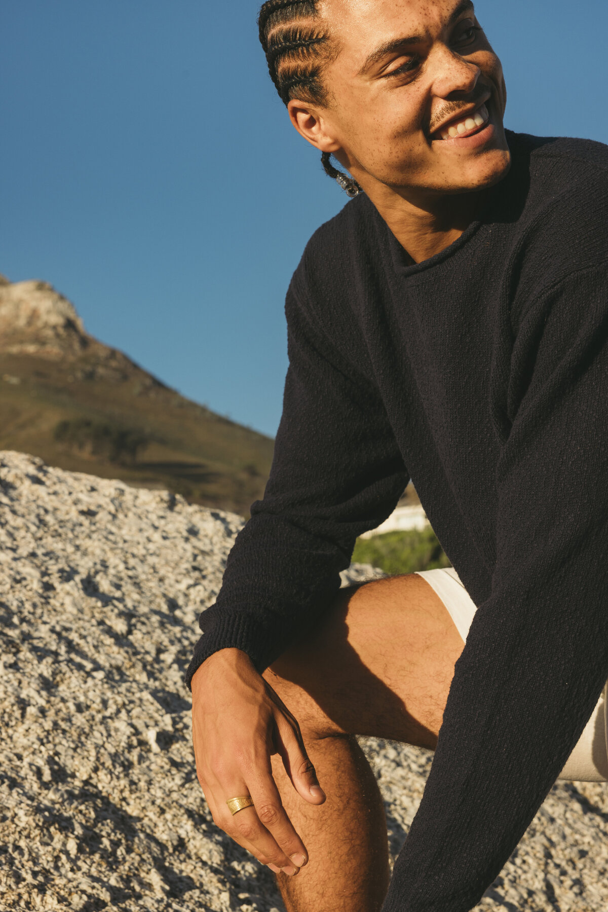 Male model Timothe smiling while wearing a dark sweater and white shorts, sitting on a rock with a mountain landscape in the background in Cape Town.