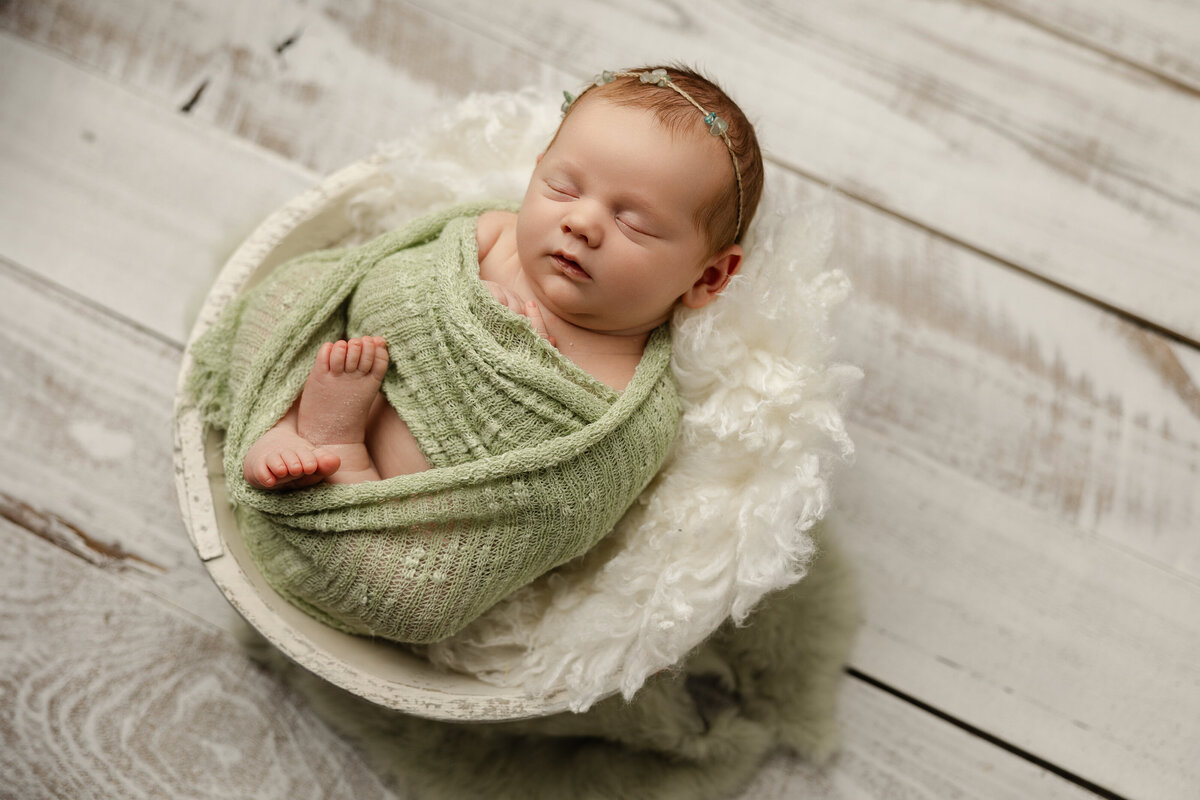 Newborn baby girl swaddled in soft green and posed in a round white bowl with textured layers on rustic wood flooring. Captured by Inland Empire newborn photographer Kaitlyn Dawn Photography.