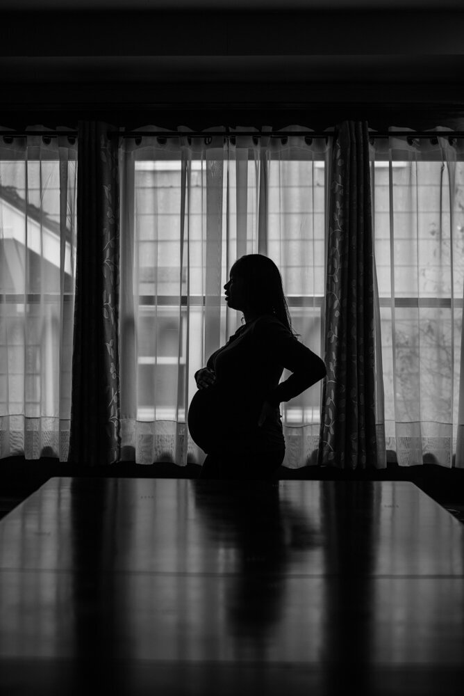 Maternity silhouette with mom resting one hand on her waist and the other on top of her belly in front of window in the dining room for home maternity session