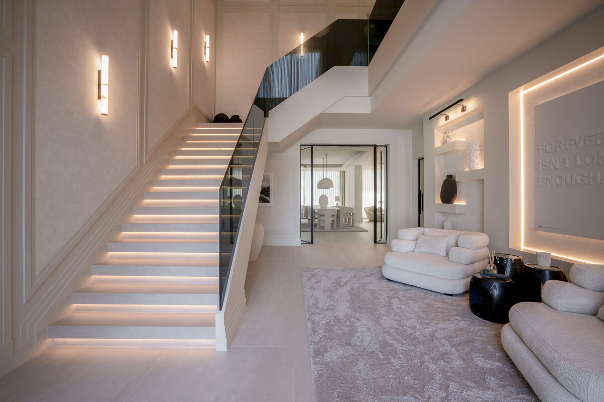 A spacious, modern entryway with a focus on light, neutral tones. A wide staircase with integrated lighting and a black glass railing ascends to the left. The walls feature vertical, cylindrical sconces emitting a warm glow. To the right, there are two plush, light-colored armchairs and a soft, textured rug. Built-in shelves with decorative items and a wall with the illuminated text 'FOREVER ISN'T LONG ENOUGH' add interest to the space. In the background, a dining area with a large table and pendant lights is visible through glass doors.