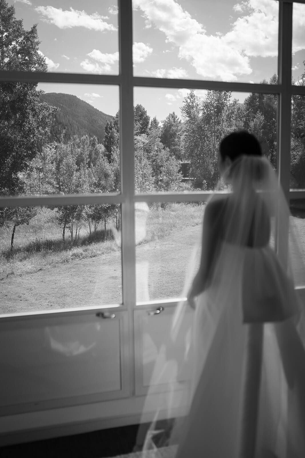 Bride gazes thoughtfully out a window at the beautiful lush summer scenery outside of the Aspen Meadows Resort wedding venue.