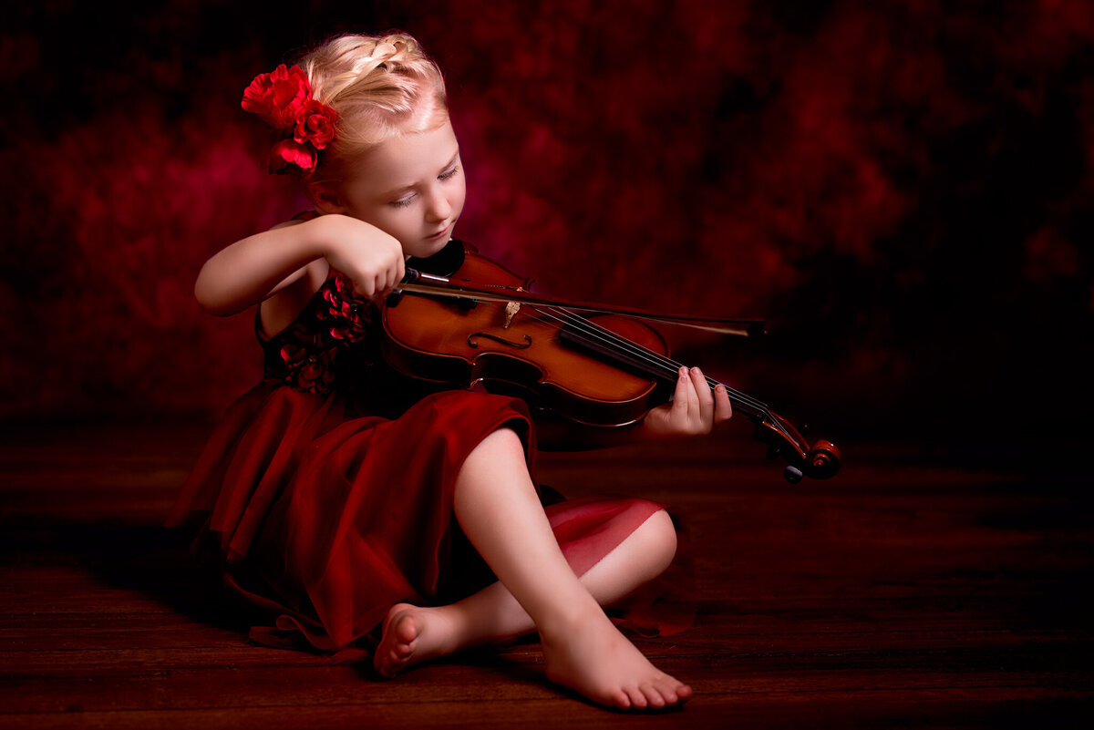 Elegant fine art portrait of a young girl holding a violin, dressed in a red dress against a dramatic dark background. Captured in Overland Park, Kansas, this artistic setup is perfect for families seeking creative and musical-themed child photography