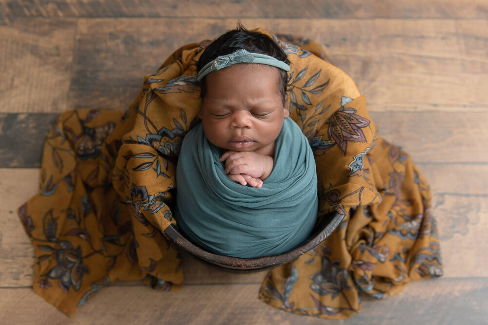 Black newborn baby girl wrapped in green in a bucket for her Hamilton, Ontario newborn photography session.