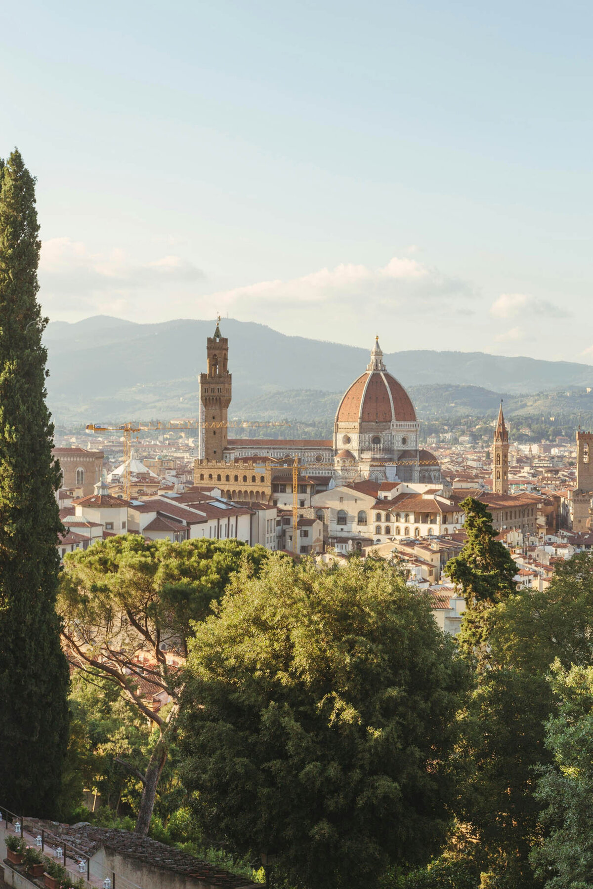 Panoramic view of Florence, Italy, featuring the iconic Duomo with its red-tiled dome. This luxury vacation scene includes historic buildings, towers, and lush greenery in the foreground, set against a backdrop of distant mountains under a clear sky.