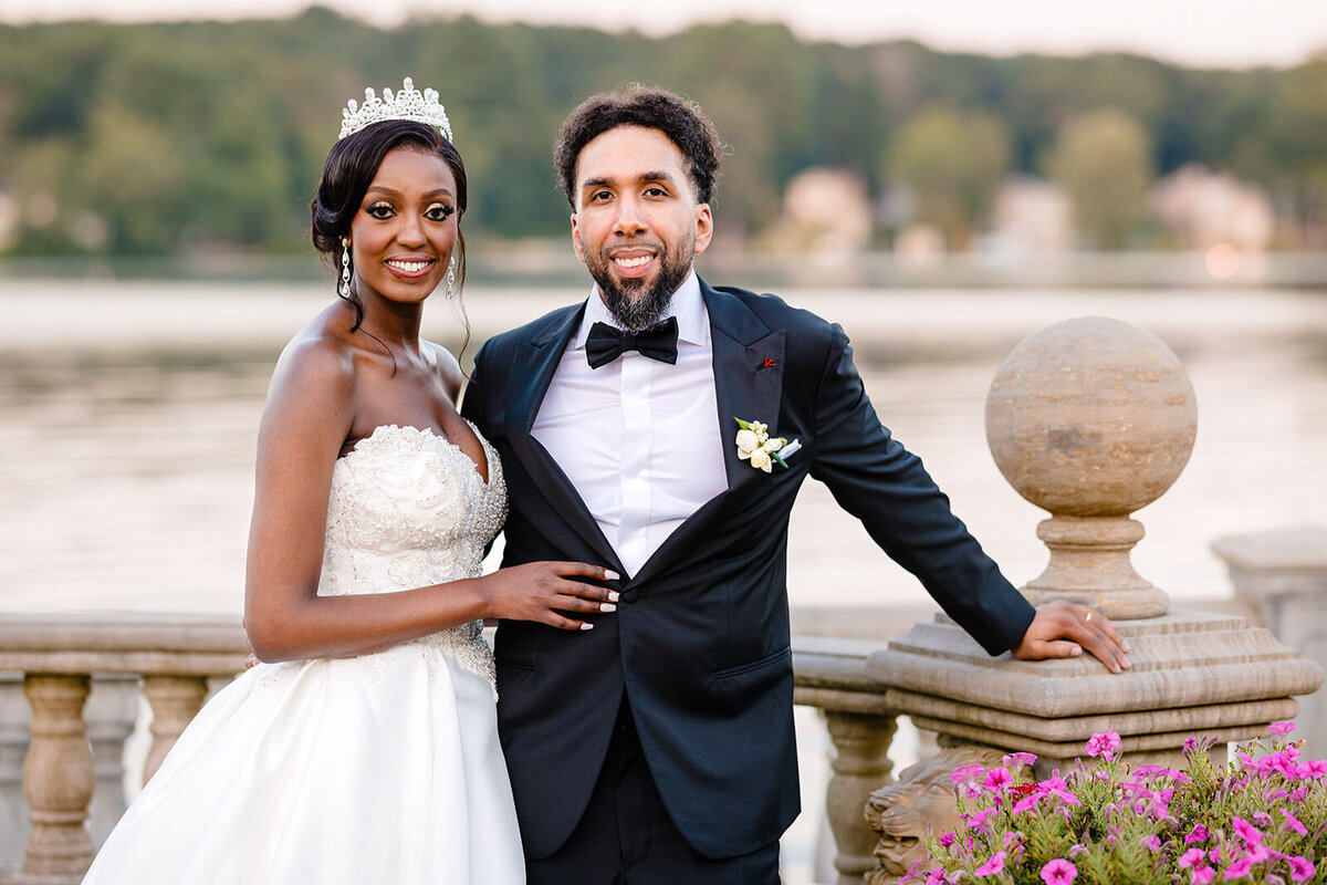 Bride and groom posing by a balustrade overlooking a lake, with the bride in a tiara and the groom in a black tuxedo