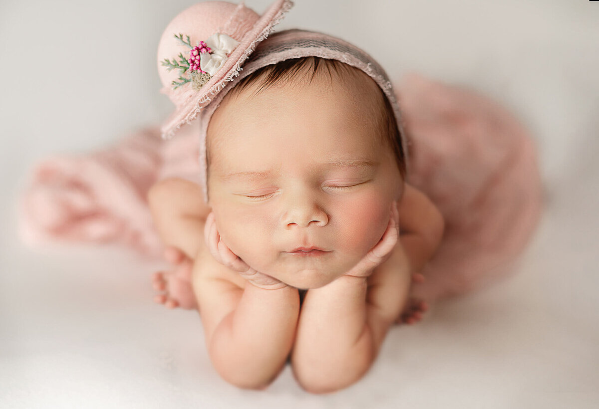 Newborn girl in a pink hat and wrapped in the froggy pose with her hands on her cheeks at her Toronto newborn photography studio session.