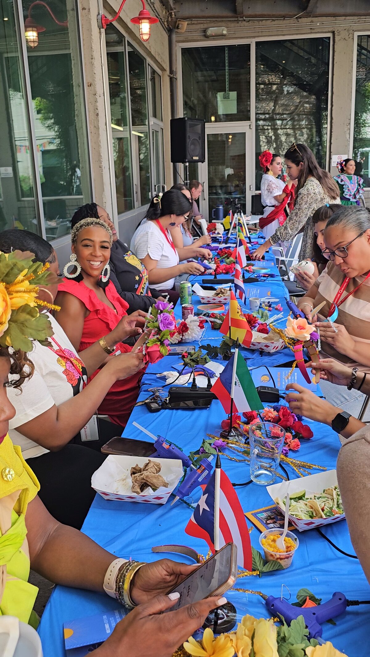 The lively scene at Soleil Vida Studio, where a group of women comes together around a giant table adorned with a blue tablecloth. Laughter and creativity fill the air as they craft vibrant flower wreaths, using a plethora of materials.