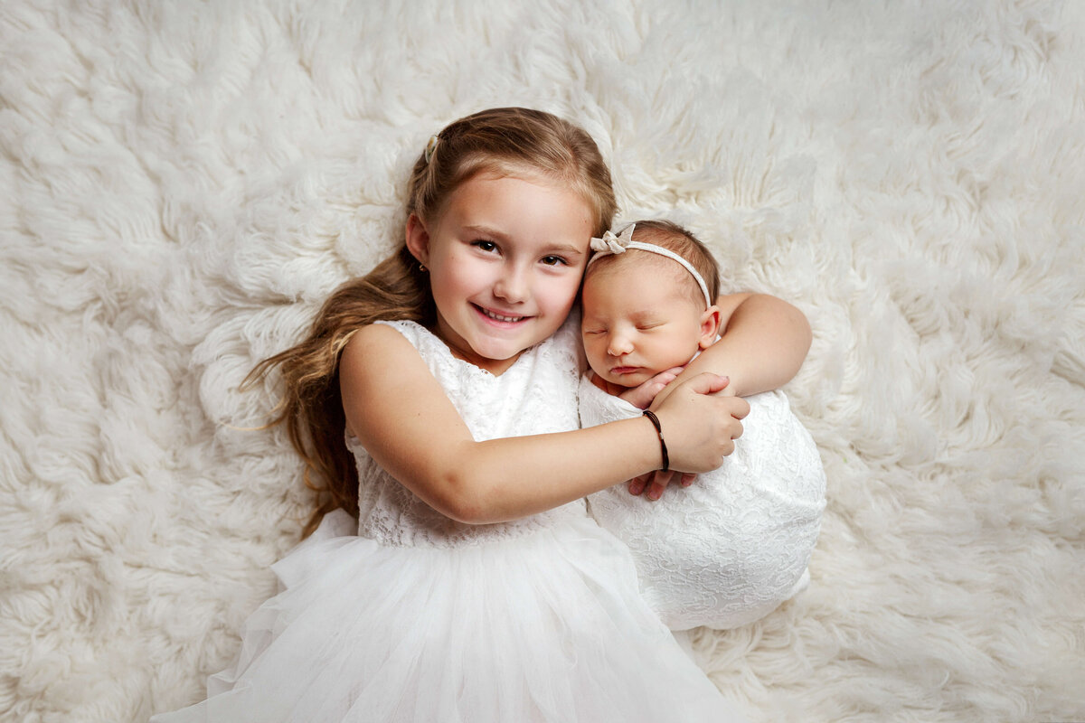 big sister wearing a pretty white dress hugging her newborn baby sister on a white fur rug in a newborn session