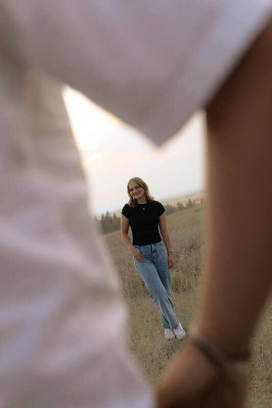 A woman stands smiling at the camera. In the foreground is a man with a hand in his pocket. You can see the woman in the negative space.