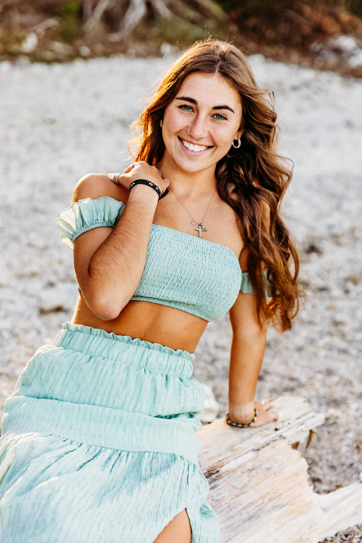 beautiful teen girl sitting on a piece of driftwood in a flowy two piece dress and smiling on a beach near Oshkosh