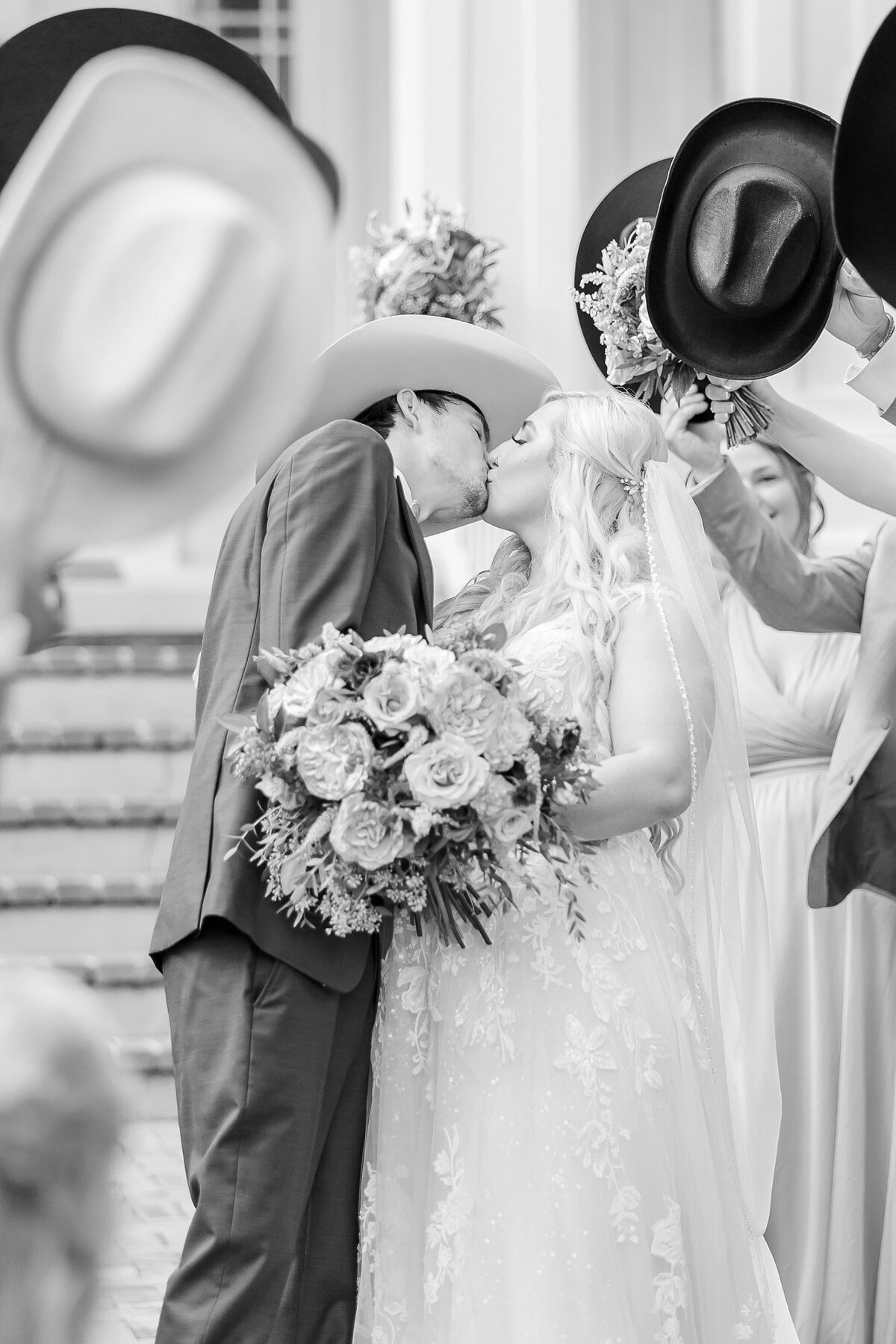 Black and white image of bride and groom kissing, surrounded by cowboy hats and flowers  | Elms of Coosada Wedding Photographer 
