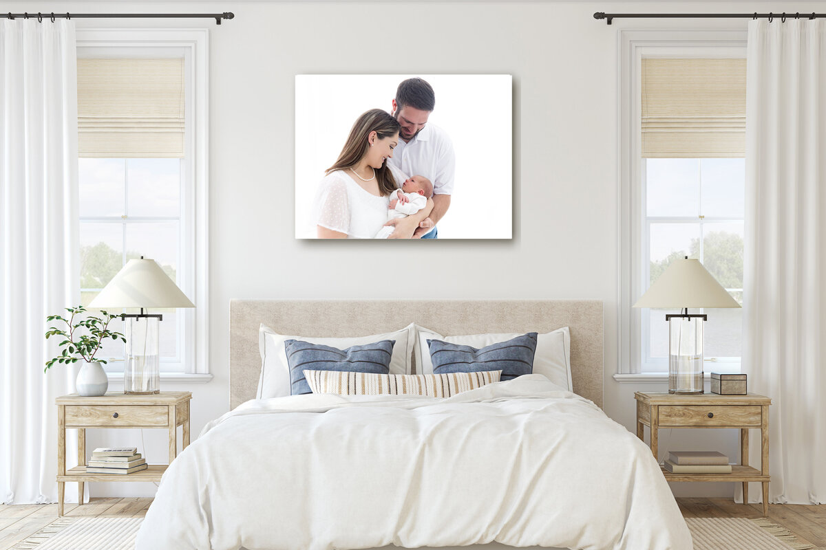 A framed fine art newborn photography portrait hangs on a wall above a bed