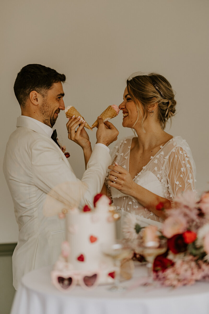 Mariés jouant à toucher le nez de leur partenaires avec des cônes de glace. Table surmontée de lunettes coeur, de wedding cake et d'un bouquet, floue, au premier plan. Séance photo pour le workshop photographie de mariage.