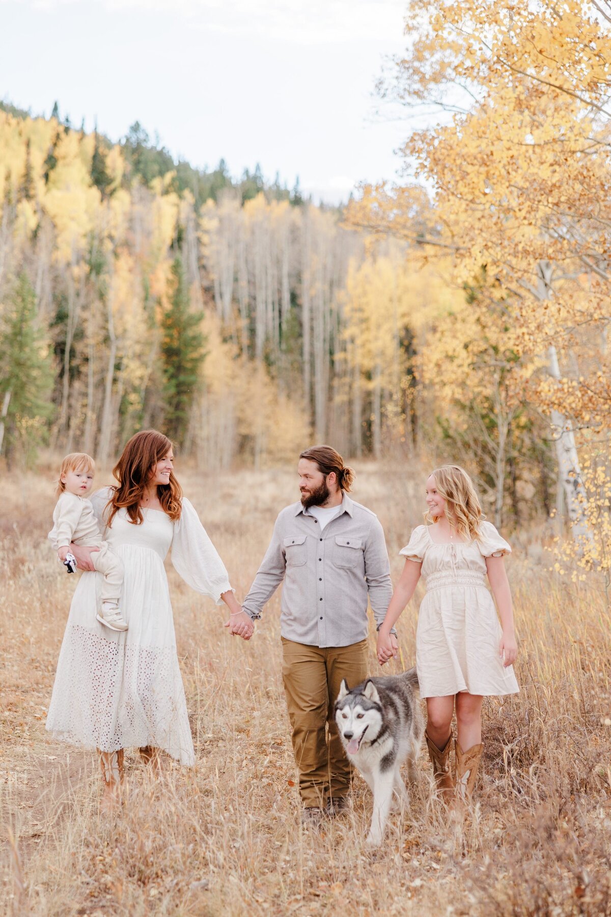 A family of 4 hold hands on a hike behind their husky in fall