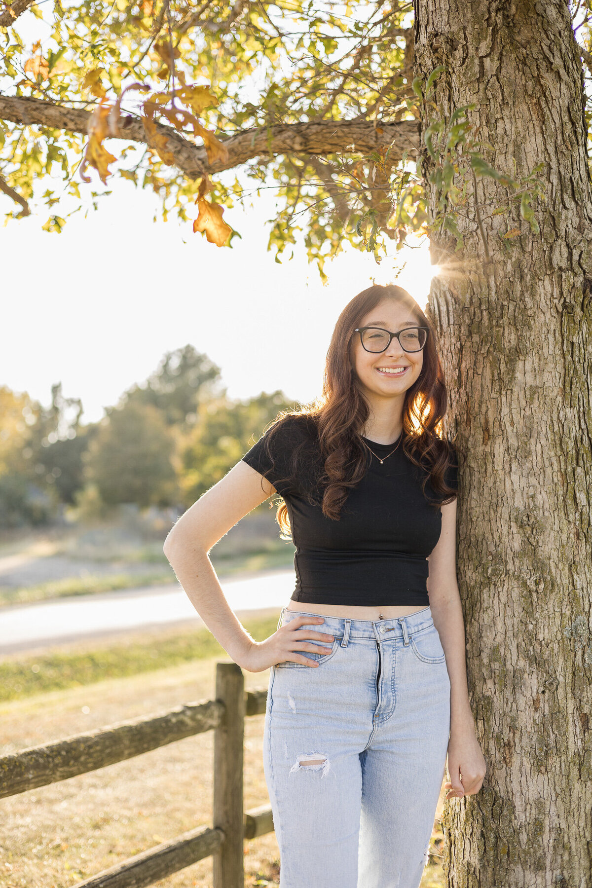 High school senior female with long dark hair, standing against a tree, with trees in the background, sun behind her, she's wearing glasses, a black short sleeve shirt, blue jeans, her hand on her hip, looking away from the camera smiling