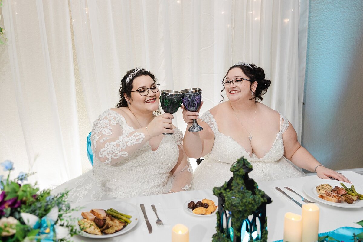 Two brides clinking fancy goblets together in a toast during their wedding reception at the A and M Gardens in Azle, Texas. The bride on the left is wearing a long sleeve, intricate, long, white dress with a tiara. The bride on the right is wearing a short sleeve, intricate, white dress also with a tiara. Their reception dinners can be seen on their head table.