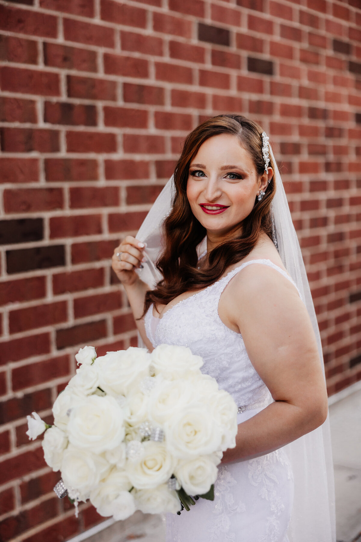 A bride holds her veil for a portrait in front of  brick wall.