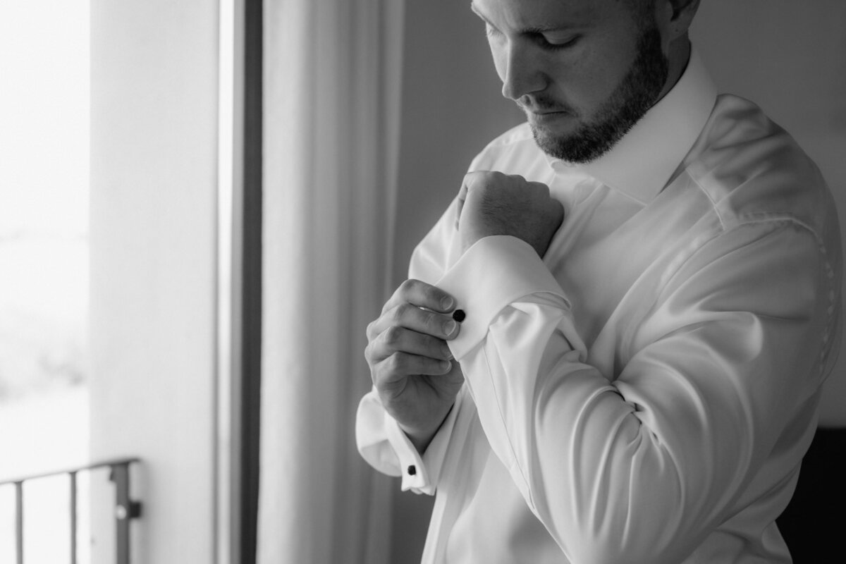 Beim Getting Ready schließt der Bräutigam seine Manschettenknöpfe vor einem Fenster stehend.