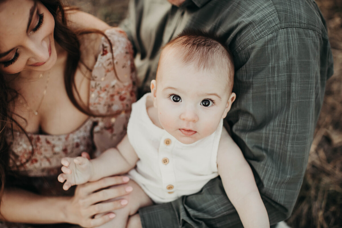 Close up of mom and dad holding baby, while baby looks up at the camera. 