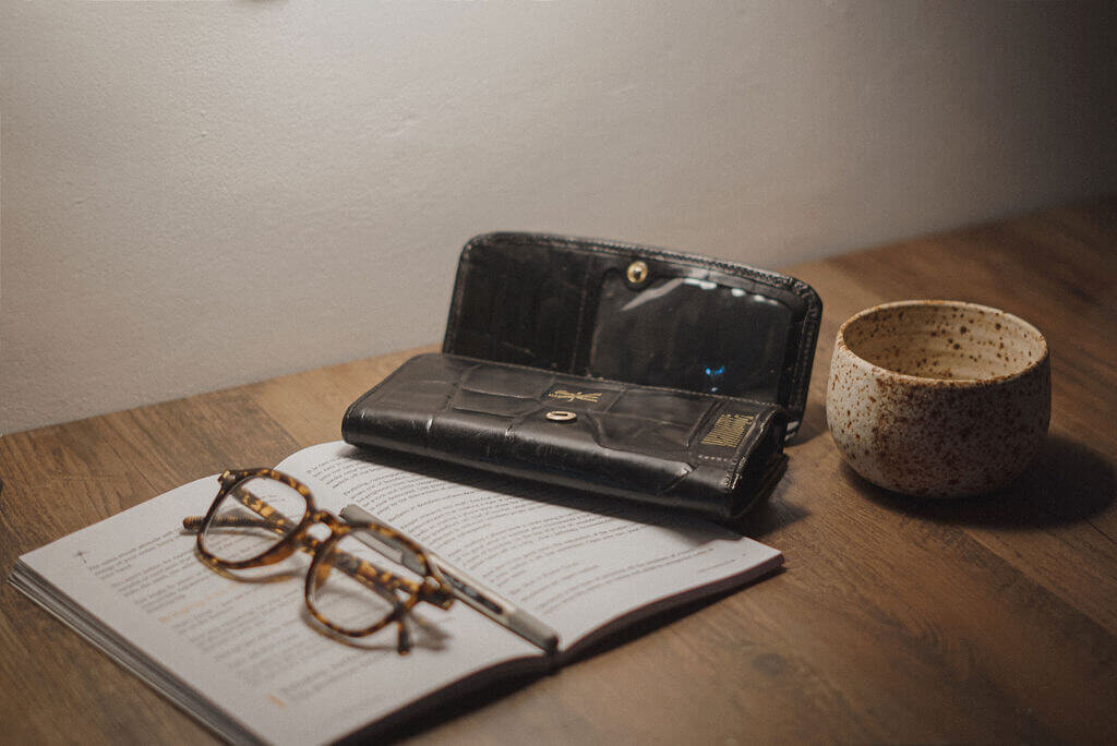 A pair of eyeglasses, an open book, a black wallet, and a ceramic cup are arranged on a wooden table under soft lighting—perfect inspiration for any brand strategist or creative from Sweet Pea Ink Creative.