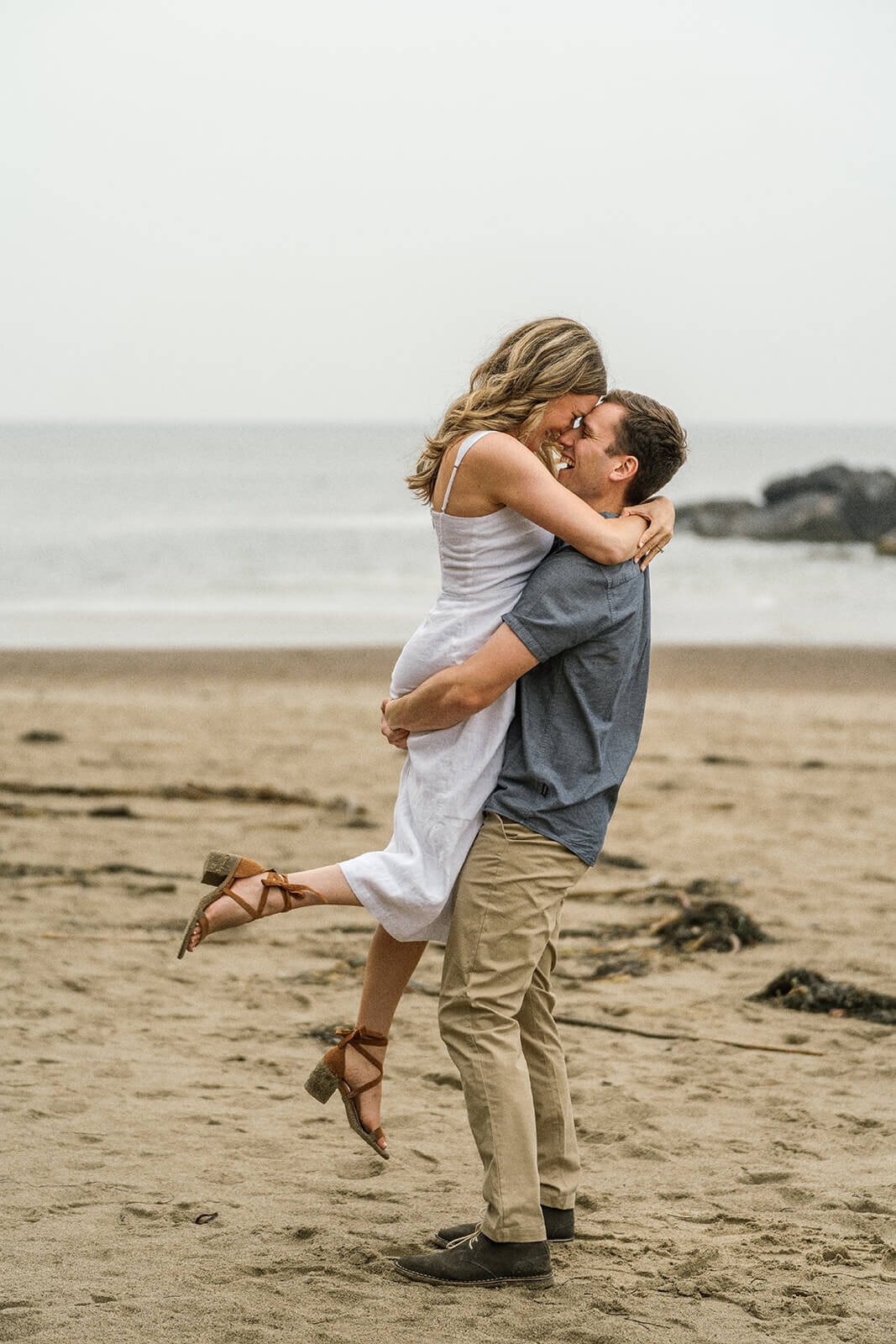 A man lifting a woman in her arms as they embrace on the bach. 