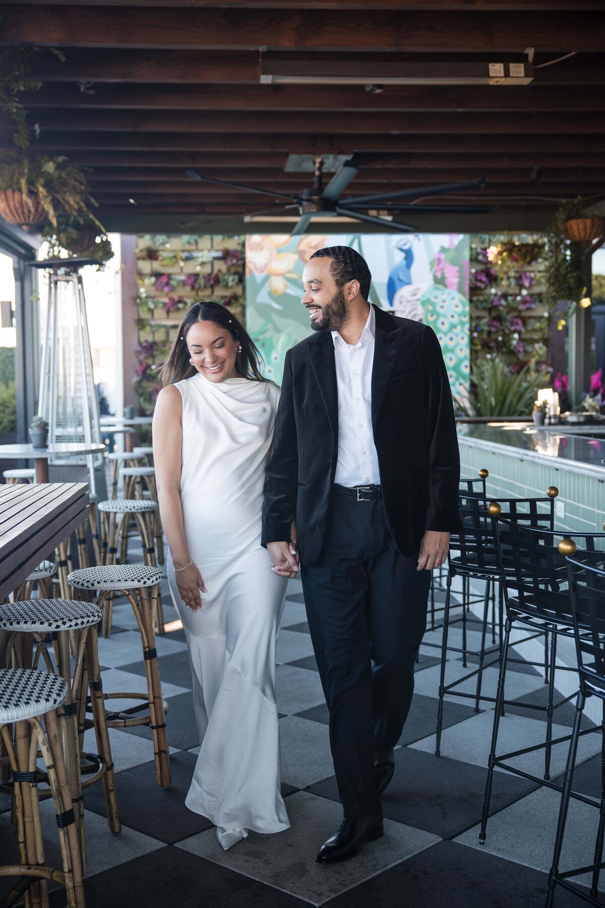A joyful couple walks hand-in-hand during their rooftop engagement session at a hotel bar, captured by Phavy Photography. The bride-to-be wears a sleek white satin gown with pointed heels, while the groom wears a black velvet blazer and dress pants. The vibrant floral mural and modern bar seating add to the upscale, editorial feel. Captured in a documentary style by a luxury wedding photographer.