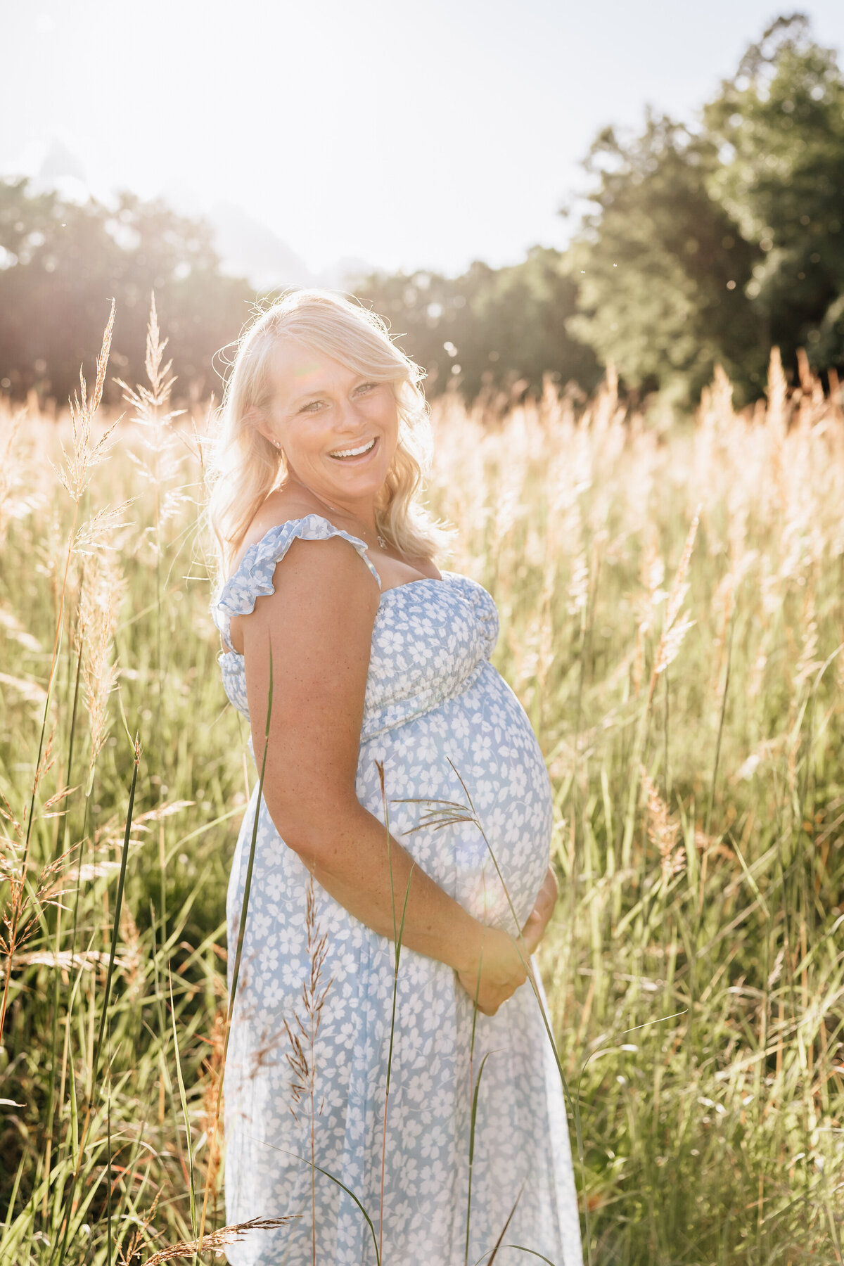 A pregnant woman smiles in a field, holding her belly.