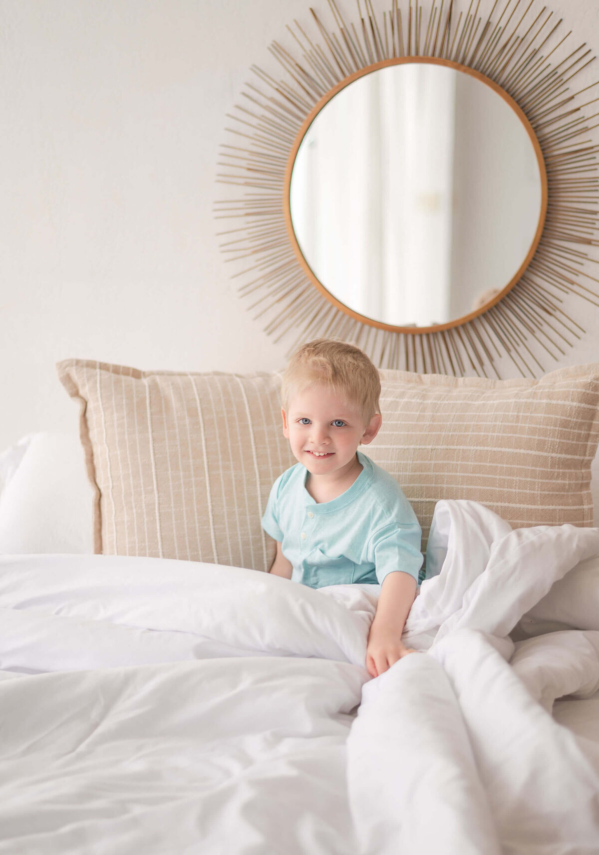 A young boy sitting on a cozy bed smiling for the camera during a relaxed in-home style studio session by Pittsburgh family photographer Lens Love Photography