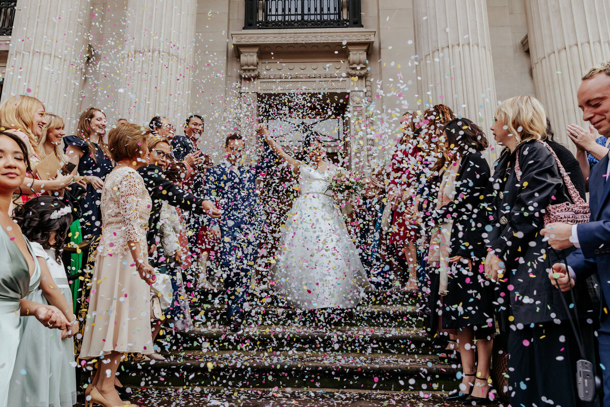 Bride and groom leave  their Old Marylebone Town Hall wedding ceremony in a flurry of confetti while holding their hands in the air