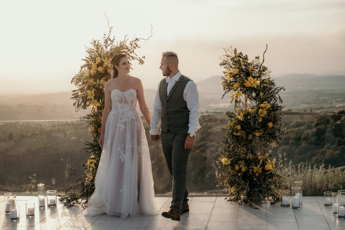 Das Hochzeitspaar steht vor zwei gelben Rosenstöcken mit der grünen Hügellandschaft der Toskana im Hintergrund.