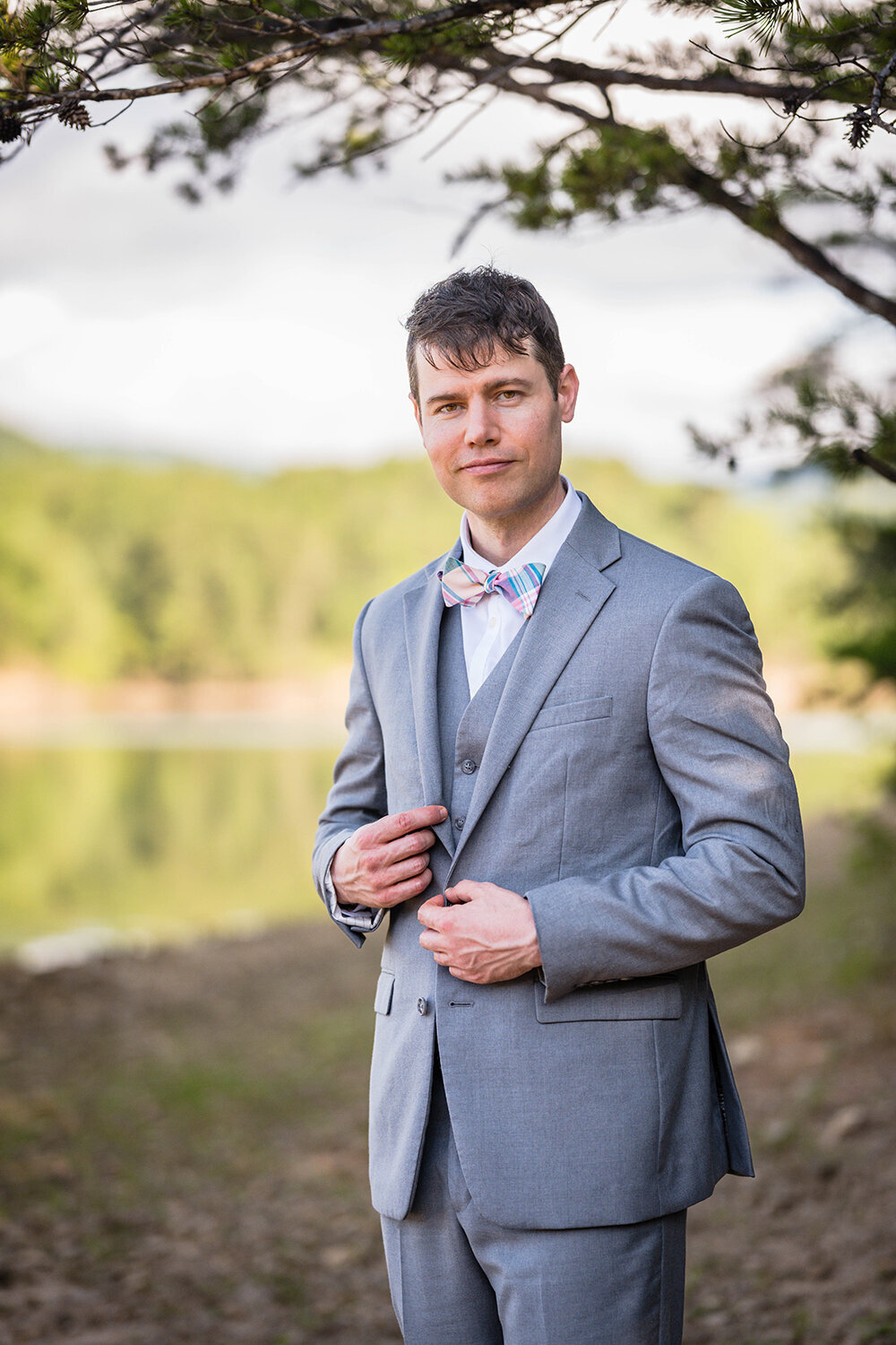 A groom stands along the shores of Carvin’s Cove for a formal portrait on his elopement day. He is wearing an all gray suit and he is grinning while grabbing onto either side of his suit jacket lapel.