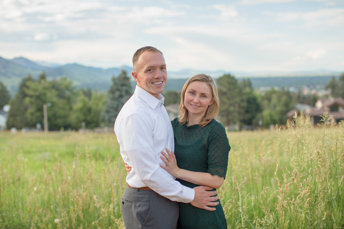 A newly engaged couple smiles in a field surrounded by mountains during their engagement session