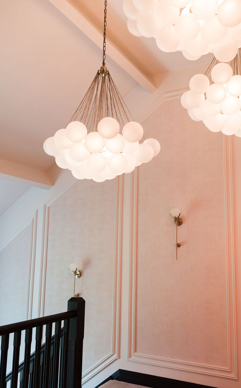 Three modern chandeliers with frosted white glass globes hang from the ceiling of a room with light-colored walls. The lights cast a warm pink glow on the walls.