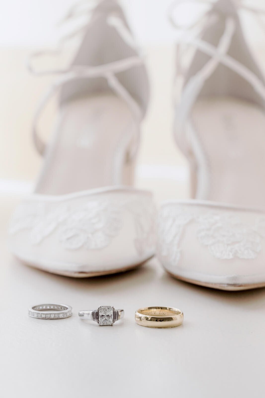 A pair of white wedding shoes, a silver ring with small diamonds, a silver ring with a diamond in the middle, and a gold ring