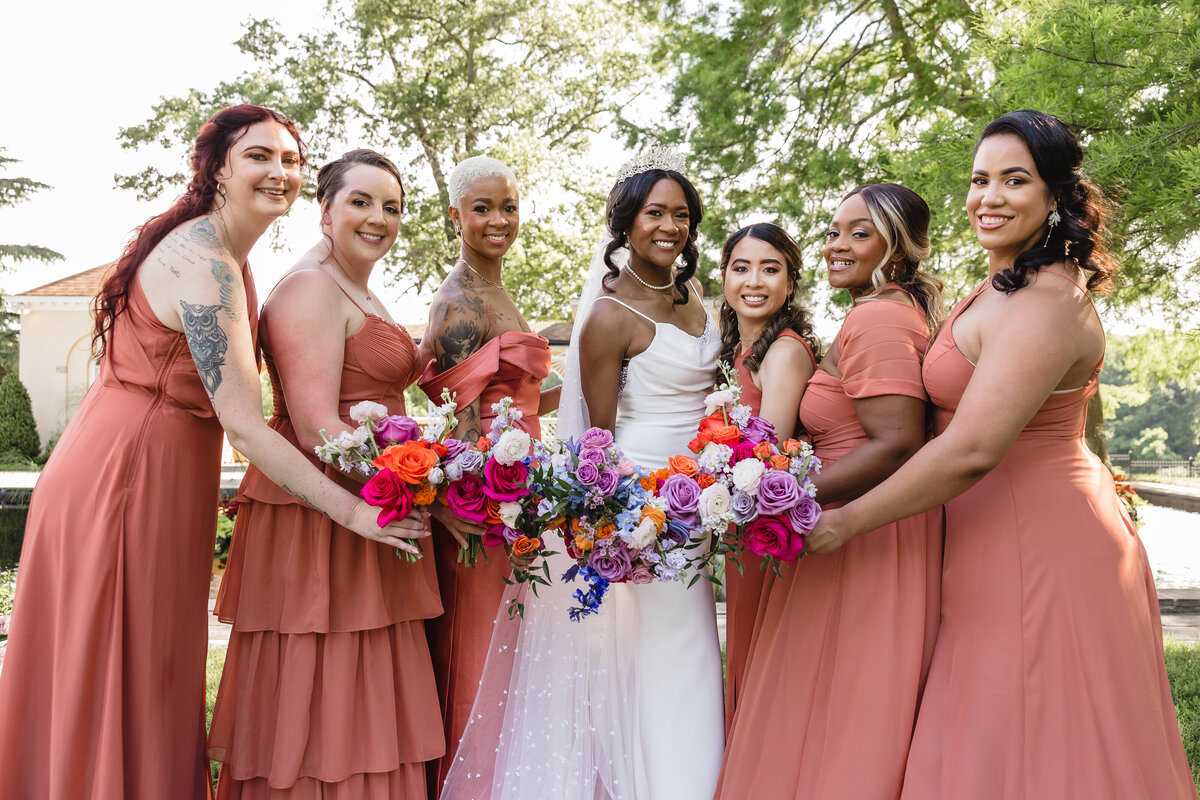 Bride holding a bouquet of pink, orange, purple and blue flowers, surrounded by bridesmaids in terracotta dresses holding matching floral arrangements.