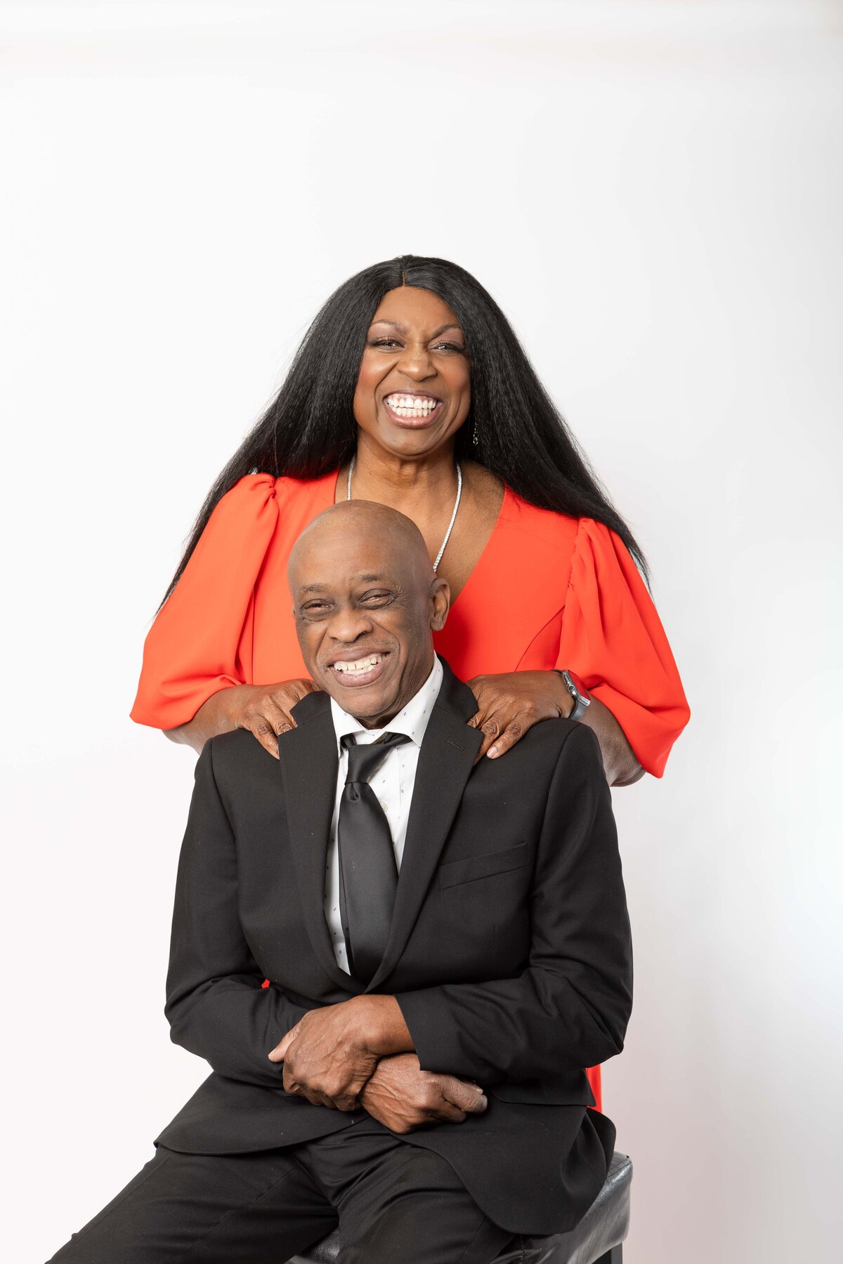 A woman in a vibrant orange dress stands behind her partner, both smiling with joy in a posed studio portrait, captured to convey warmth and authenticity for a personal brand.