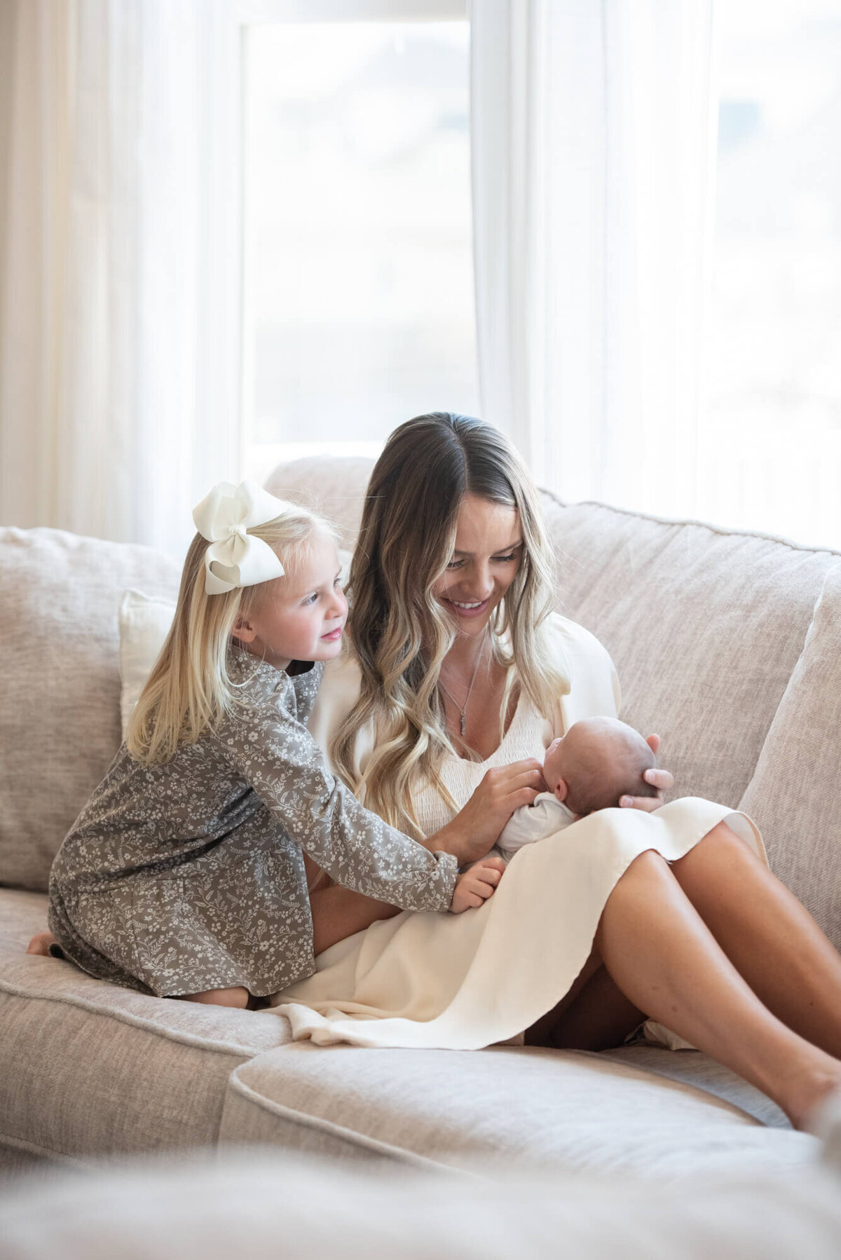 A toddler girl climbs over mom to play with newborn baby in mom's lap