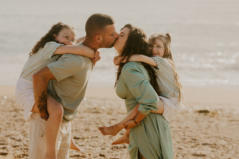 Deux parents s'embrassant et portant chacun une de leur fille sur le dos pour une séance photo en Vendée, à la plage.