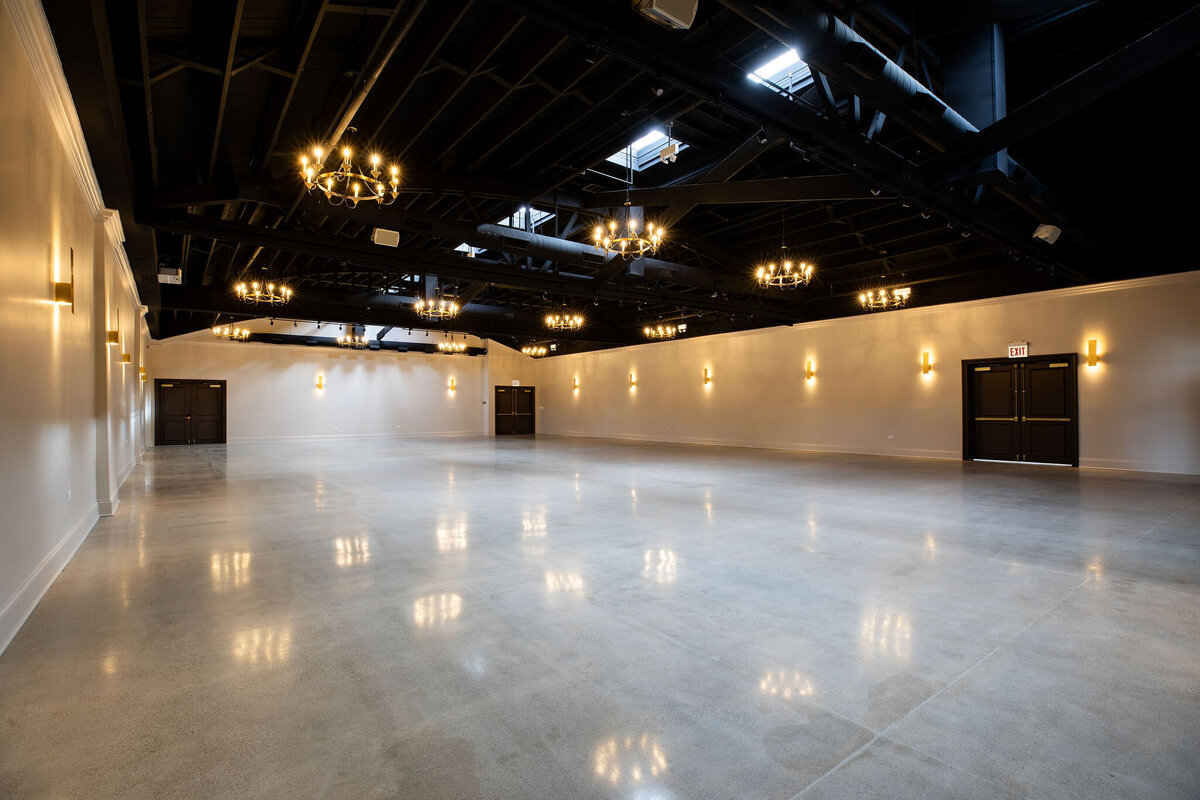 Spacious ceremony, reception or event space, with high industrial ceilings, polished floors, neutral walls and details, and ample natural light streaming through skylights and all around sconces and chandeliers, ready to be set up for an event