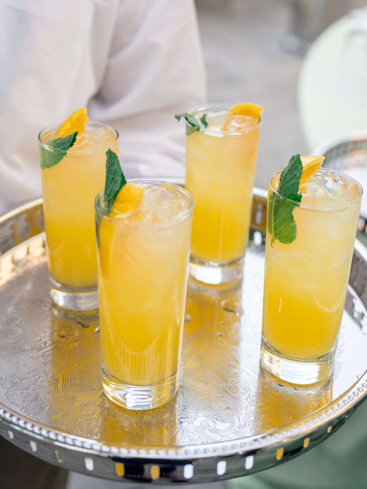 Four tall glasses of iced yellow drinks garnished with mint and lemon slices are arranged on an ornate silver tray, carried by a person in a white shirt.