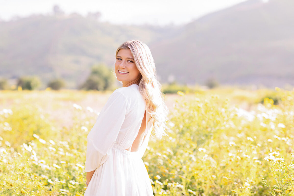 A person in a white dress standing in a field of yellow and white wildflowers, smiling at the camera. The background features soft-focus hills under a clear sky.