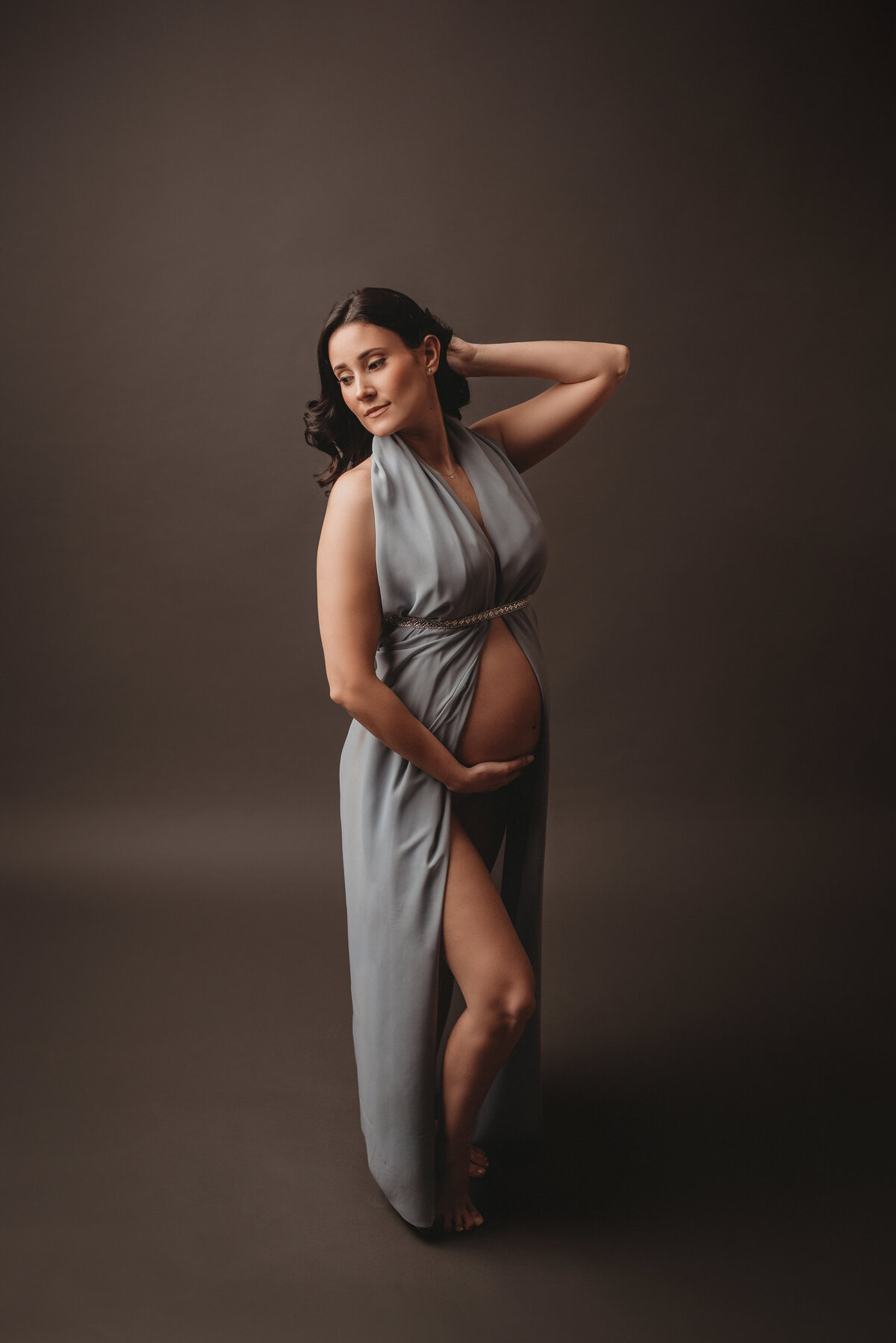 Maternity picture of goddess looking brown haired woman in light blue chiffon open tummy dress. She is holding her baby bump with right hand and other hand is tucked behind her hair while she looks over her right shoulder